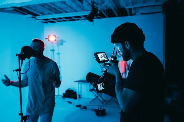 Video shoot, blue lighting, camera man holding camera recording artist on mic, in a photo studio, white walls